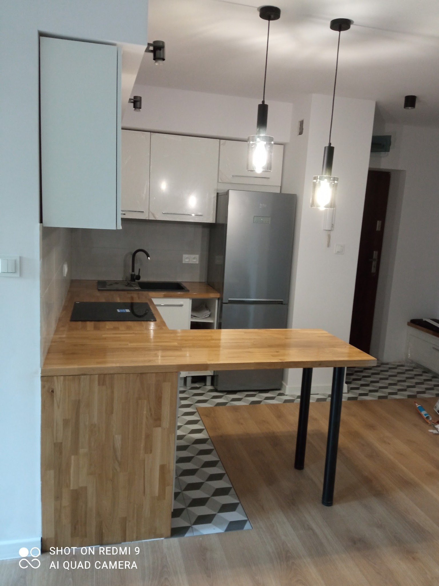 Nowoczesna kuchnia z drewnianym blatem i białymi szafkami, oświetlona wiszącymi lampami. W tle widoczna lodówka i wzorzysta podłoga, tworząca unikalny charakter wnętrza.