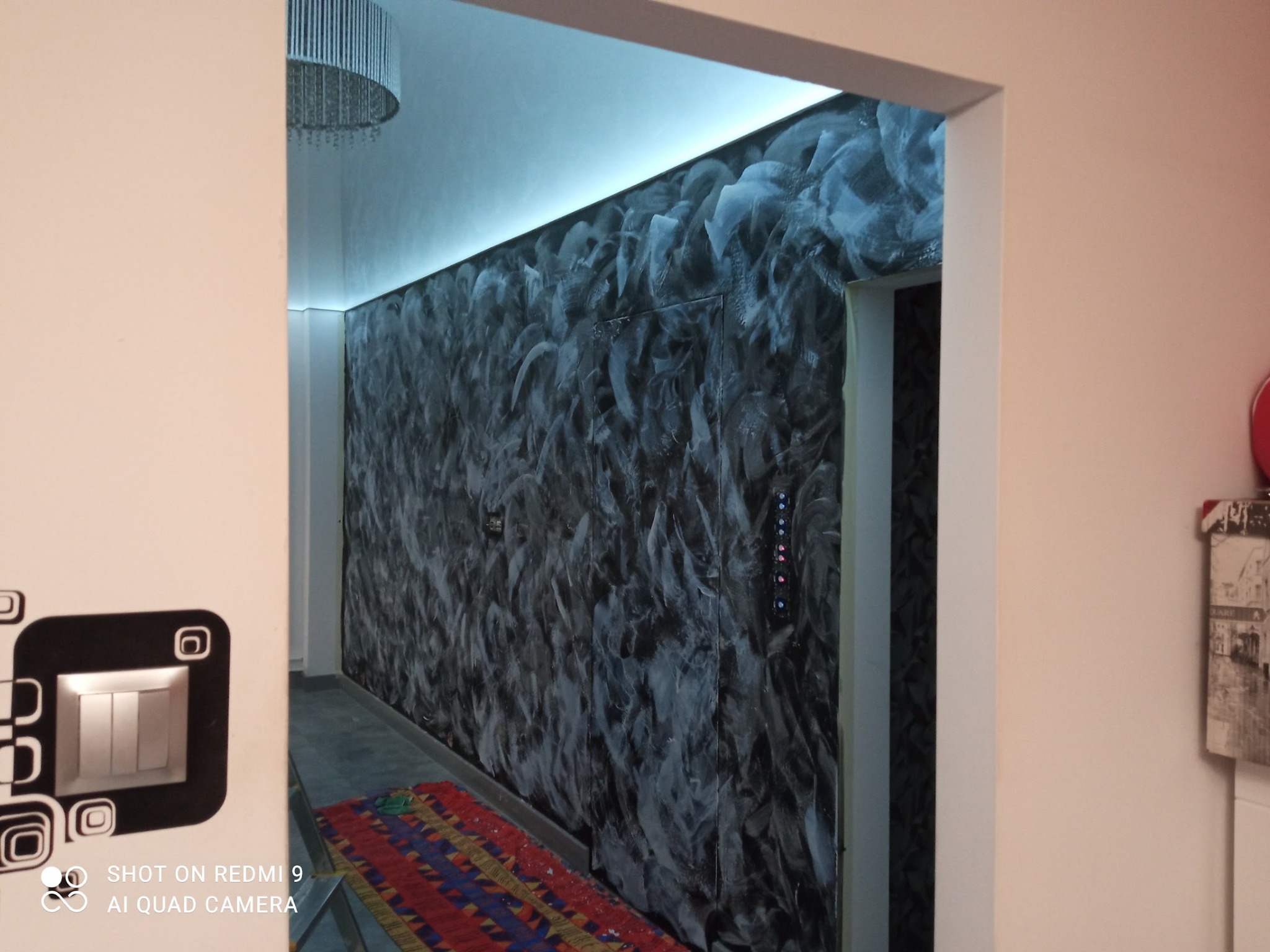 Ściana z efektem dekoracyjnym w odcieniach szarości, widoczna przez framugę drzwi. Na ścianie włącznik światła z wzorem. W tle widoczny fragment sufitu z lampą.