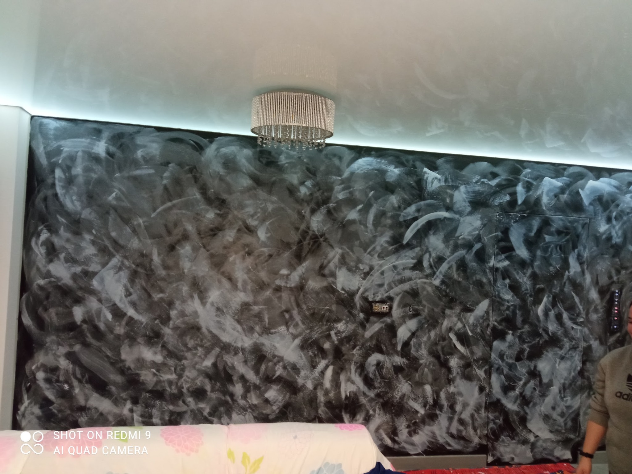 Ściana z dekoracyjną, czarno-białą fakturą, oświetlona listwą LED pod sufitem i wiszącym żyrandolem. Widoczny fragment sofy z kwiatowym wzorem.