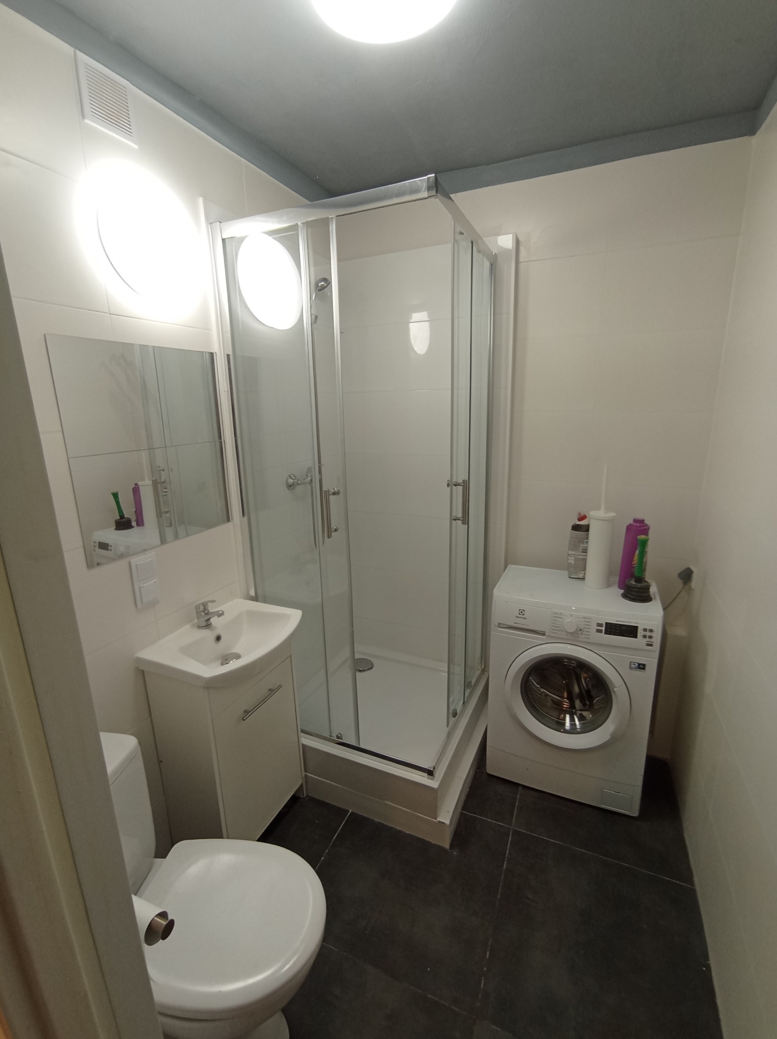 Funkcjonalna łazienka po remoncie: kabina prysznicowa, umywalka z szafką, toaleta i pralka Electrolux na ciemnej podłodze. Białe płytki na ścianach, szary sufit.