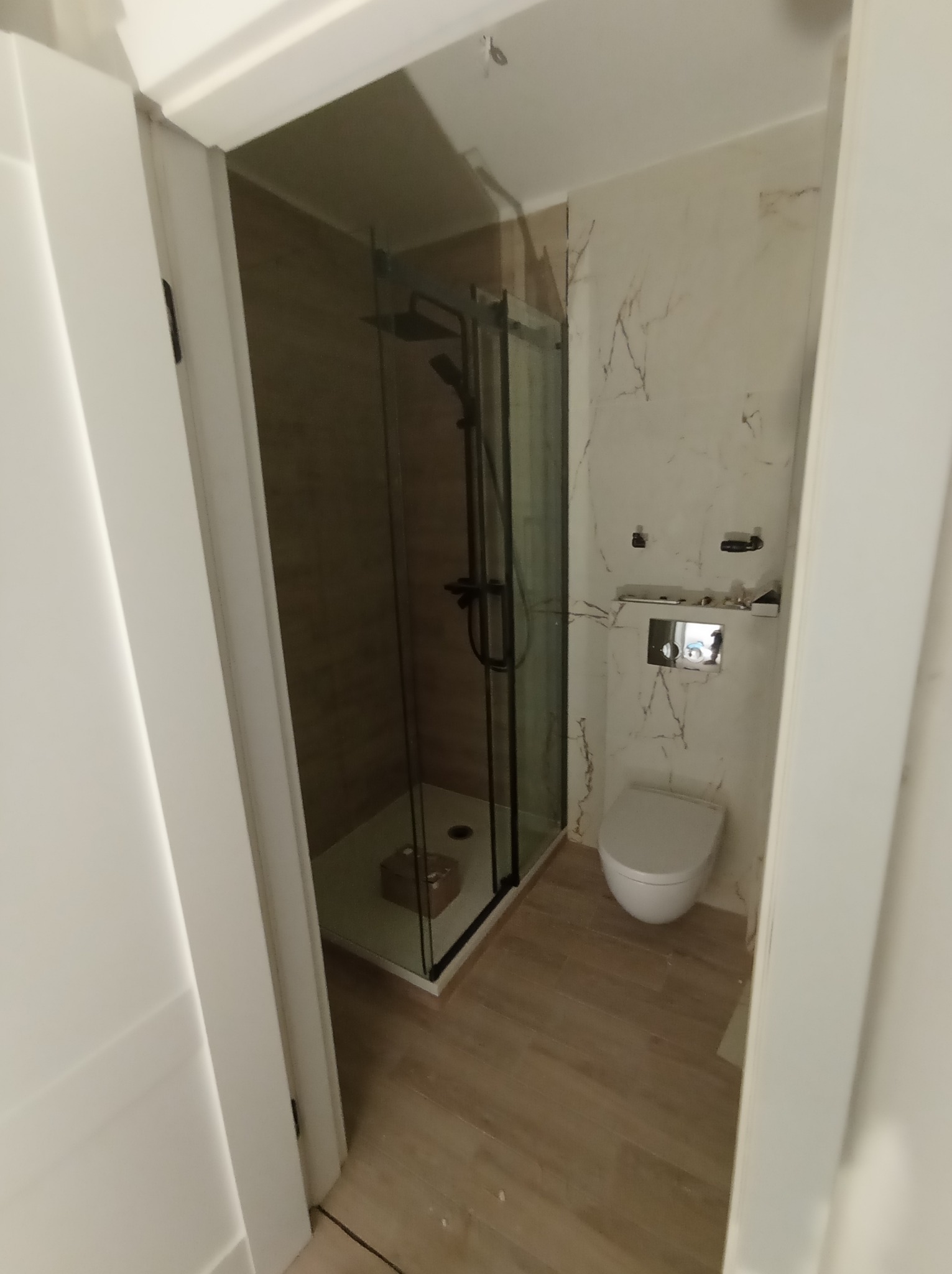 Nowoczesna łazienka z kabiną prysznicową typu walk-in z czarnymi profilami, podwieszaną toaletą i marmurowymi płytkami na ścianach. Widok zza otwartych drzwi.