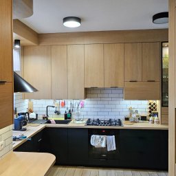 Gniazdko.lu - Nowoczesna kuchnia z jasnymi, drewnianymi szafkami górnymi i czarnymi dolnymi. Widoczny czarny okap, białe kafelki i drewniana podłoga. Oświetlenie podszafkowe.