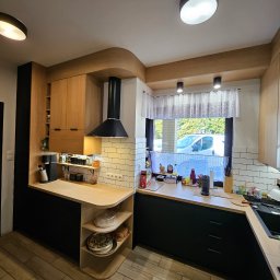 Gniazdko.lu - Nowoczesna kuchnia z czarnymi szafkami dolnymi i drewnianymi górnymi, oświetlona lampami sufitowymi. Widoczny okap i okno z firanką.