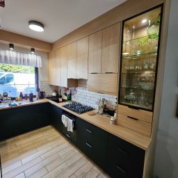 Gniazdko.lu - Nowoczesna kuchnia z jasnymi drewnianymi frontami szafek górnych i czarnymi dolnymi, z oświetloną witryną na szkło i sprzęt AGD w tle.