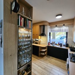 Gniazdko.lu - Nowoczesna kuchnia z drewnianymi szafkami, czarnymi blatami i witryną z kolekcją porcelany. Uwagę przykuwa zegar na ścianie i jasna podłoga.