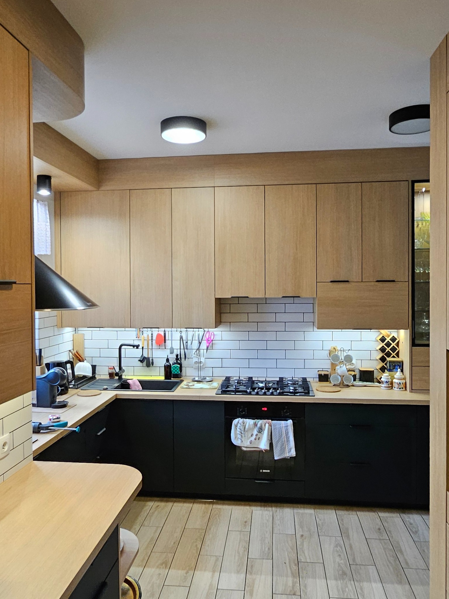 Nowoczesna kuchnia z jasnymi, drewnianymi szafkami górnymi i czarnymi dolnymi. Widoczny czarny okap, białe kafelki i drewniana podłoga. Oświetlenie podszafkowe.