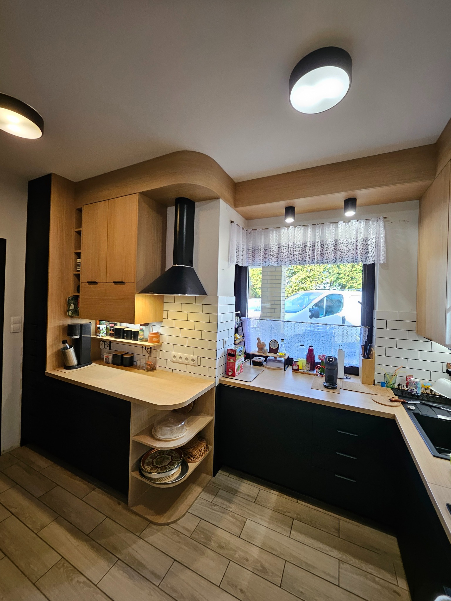 Nowoczesna kuchnia z czarnymi szafkami dolnymi i drewnianymi górnymi, oświetlona lampami sufitowymi. Widoczny okap i okno z firanką.