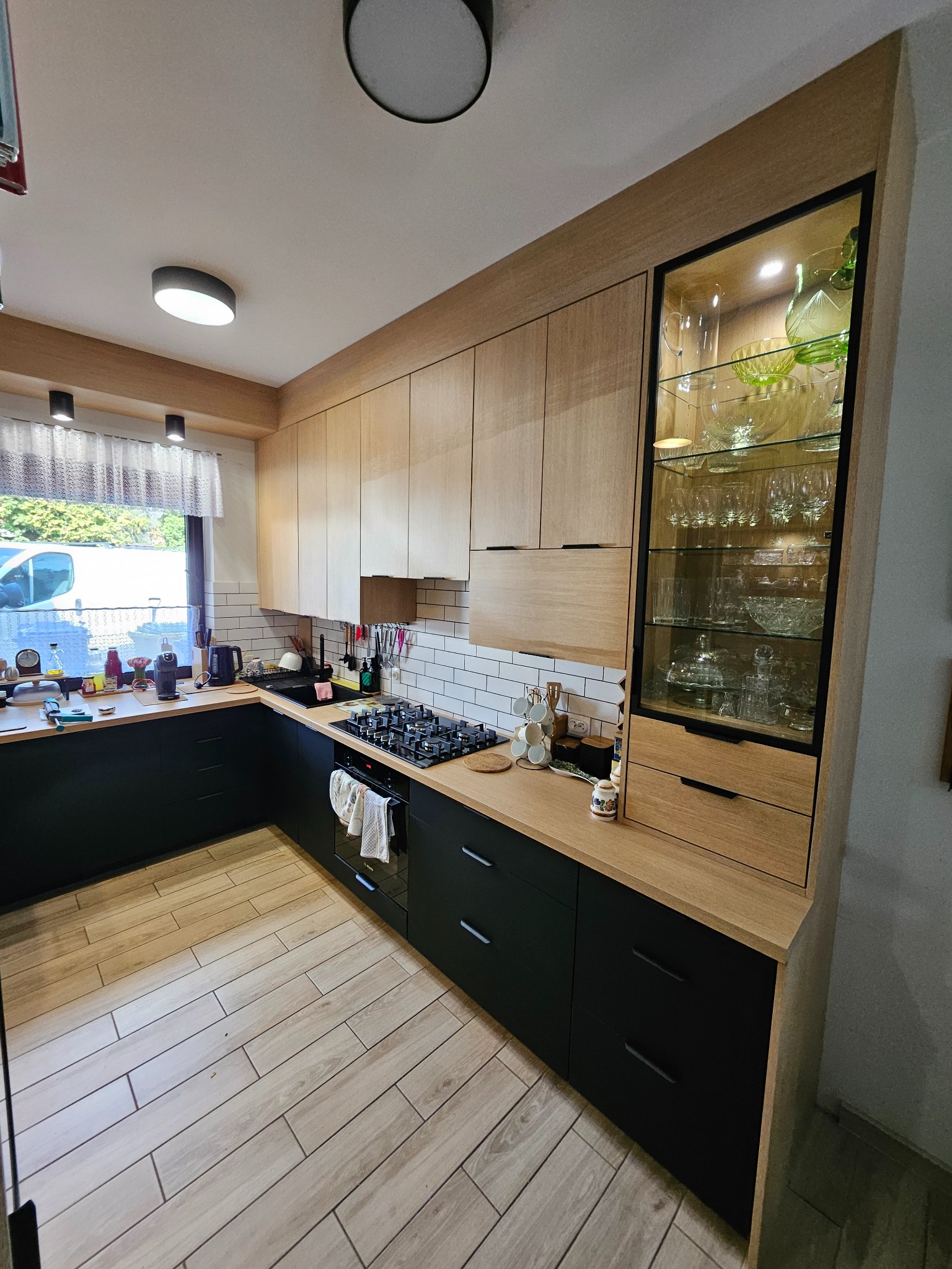 Nowoczesna kuchnia z jasnymi drewnianymi frontami szafek górnych i czarnymi dolnymi, z oświetloną witryną na szkło i sprzęt AGD w tle.