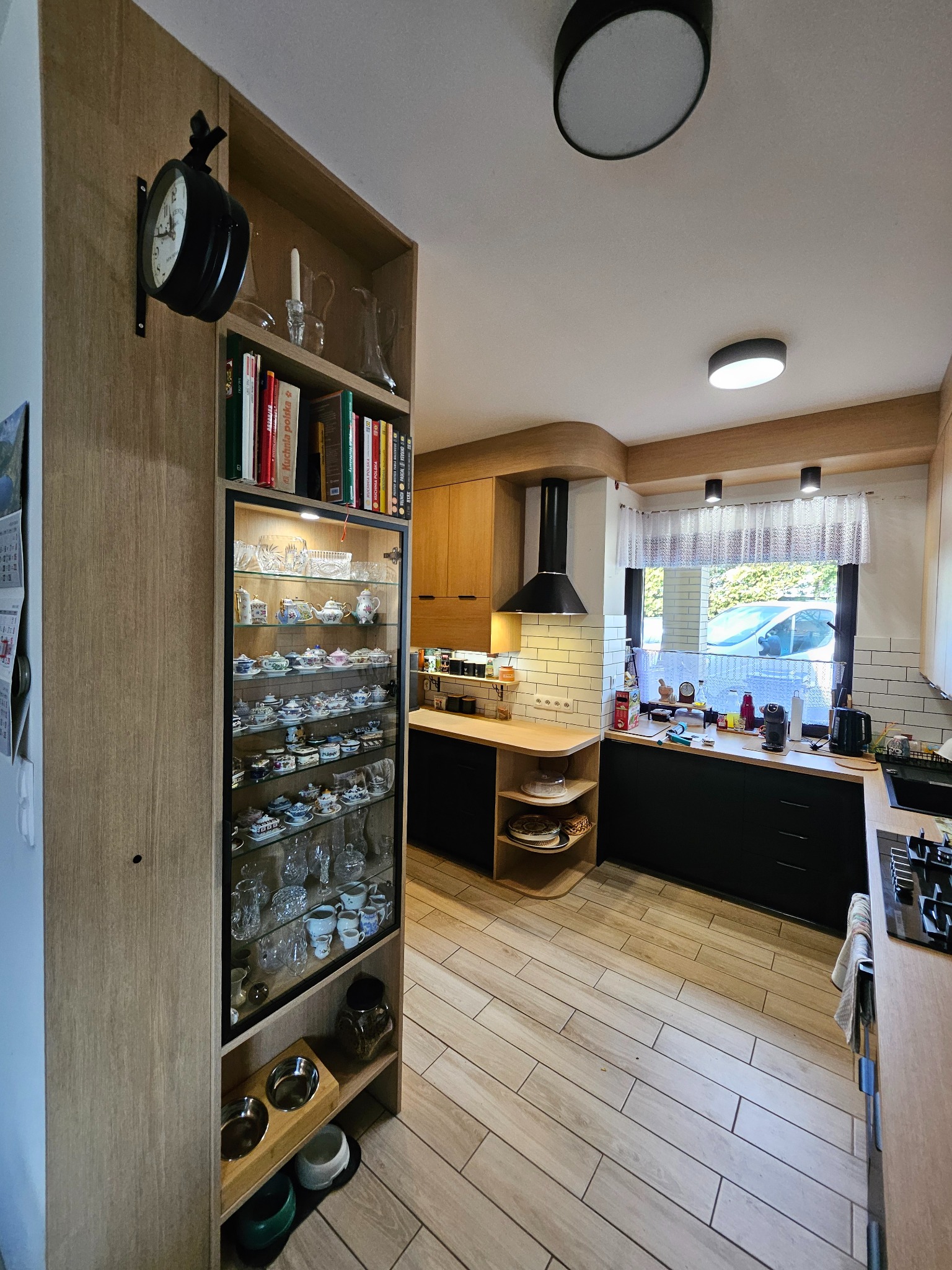 Nowoczesna kuchnia z drewnianymi szafkami, czarnymi blatami i witryną z kolekcją porcelany. Uwagę przykuwa zegar na ścianie i jasna podłoga.