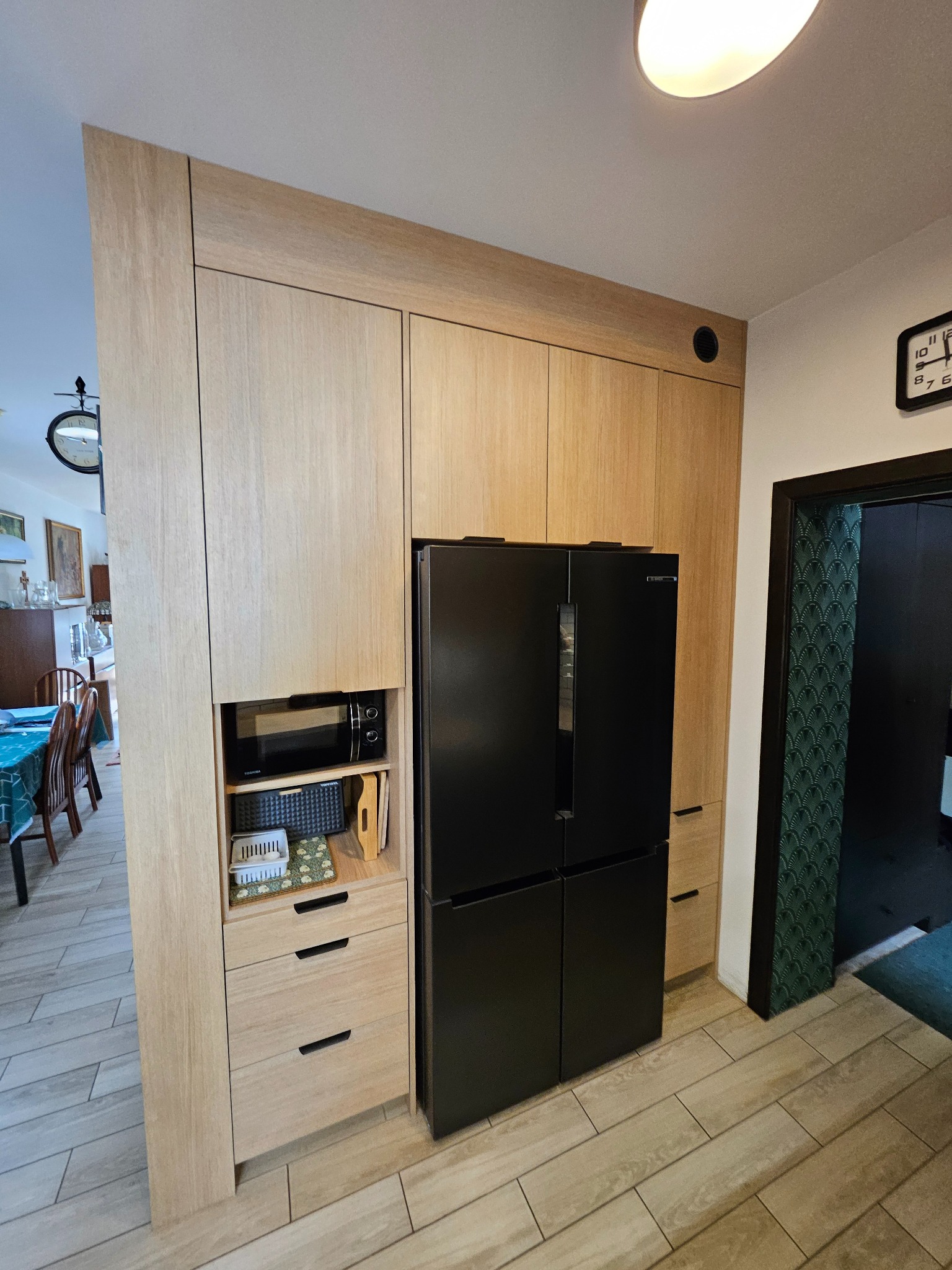 Jasna zabudowa kuchenna z drewnianymi frontami, wkomponowana czarna lodówka i mikrofalówka. Widok na jadalnię i wzorzystą tapetę w przejściu.