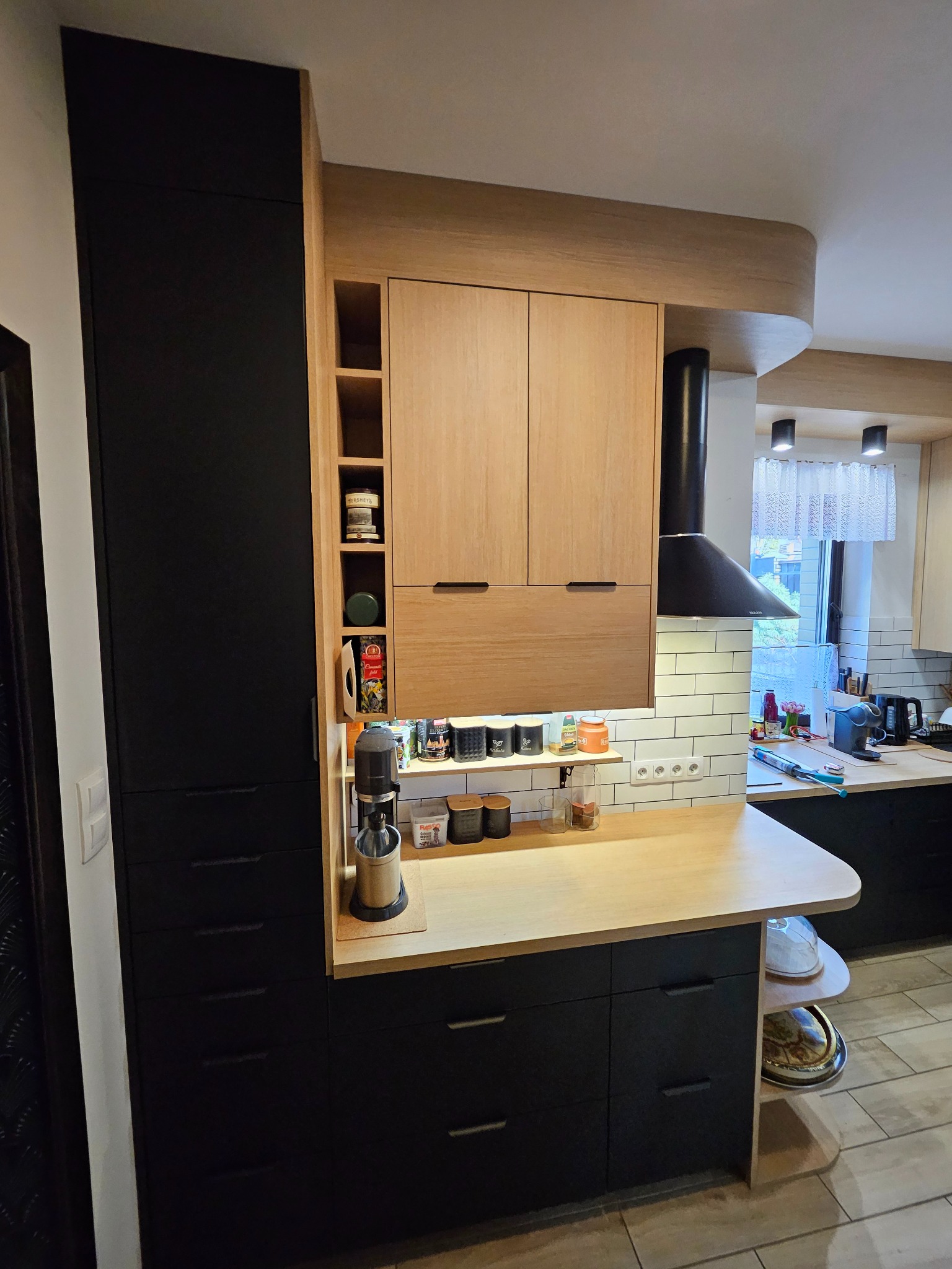 Nowoczesna kuchnia z czarnymi szafkami dolnymi i drewnianymi górnymi, z otwartymi półkami i blatem. Widoczny okap i akcesoria kuchenne. Ujęcie z perspektywy.