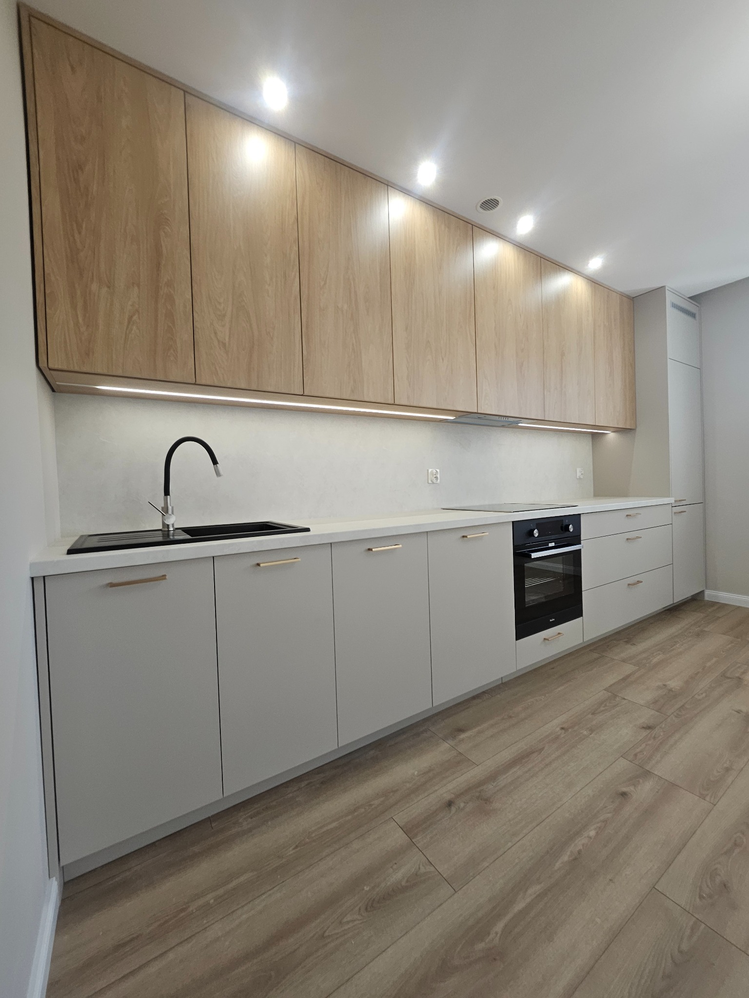 Jasna kuchnia z drewnianymi szafkami górnymi i szarymi dolnymi, czarny zlew i piekarnik, złote uchwyty, oświetlenie LED pod szafkami, parkiet. Nowoczesny design.