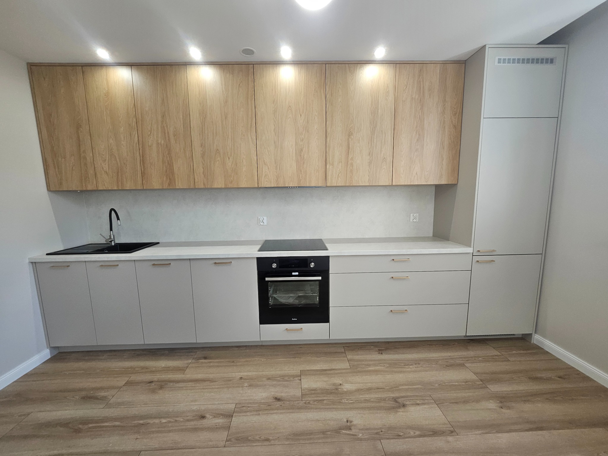 Jasna kuchnia z drewnianymi szafkami górnymi i szarymi dolnymi, z czarnym zlewem i płytą. Podłoga z paneli imitujących drewno. Oświetlenie punktowe.