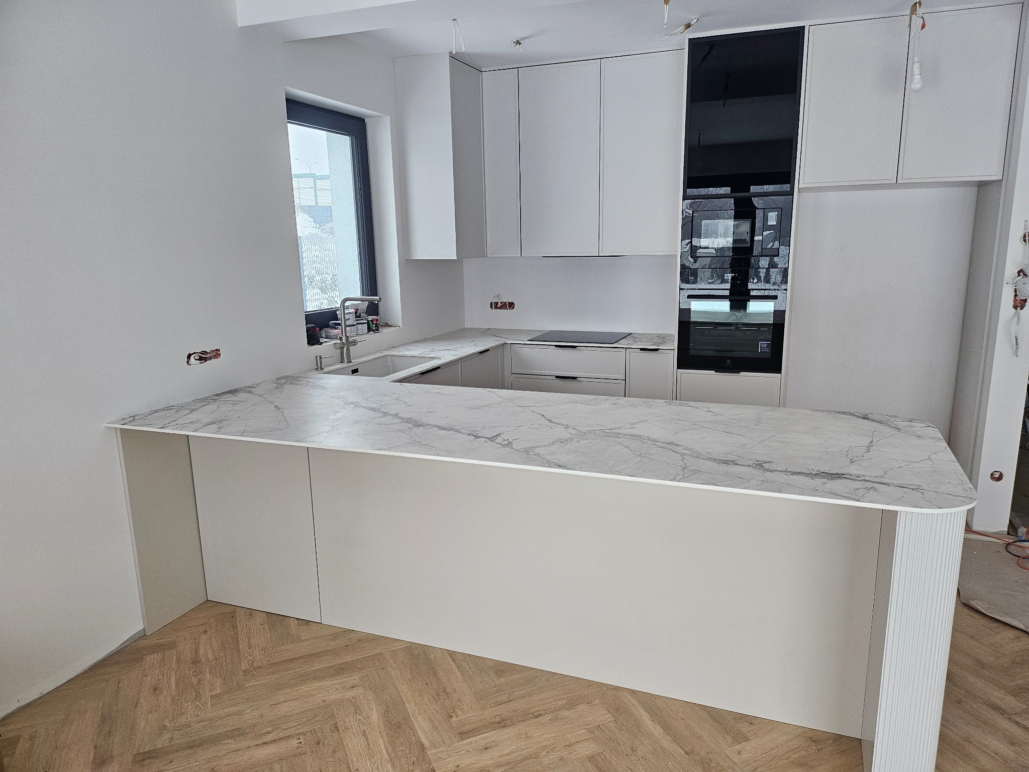 Nowoczesna, jasna kuchnia z wyspą i marmurowym blatem. Minimalistyczne szafki, czarne akcenty. Widoczne niedokończone wykończenia ścian.