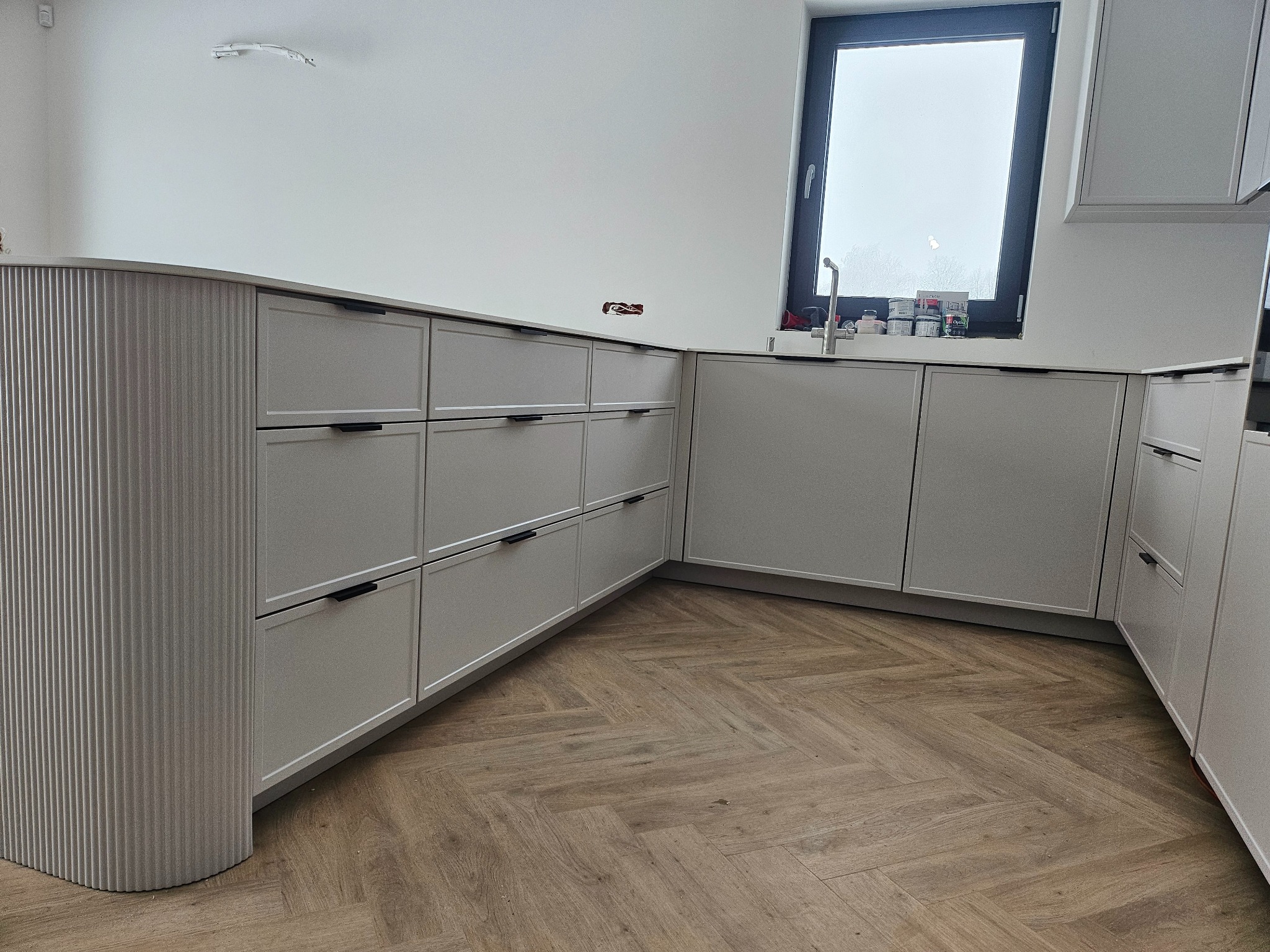 Szara zabudowa kuchenna z frezowanym frontem szafki narożnej, czarne uchwyty, drewniana podłoga w jodełkę, okno z widokiem na zewnątrz. Minimalistyczny design.