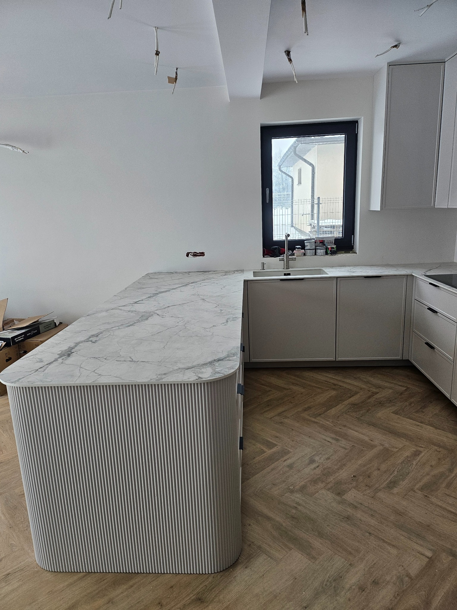 Jasna kuchnia z marmurowym blatem i ryflowanym frontem wyspy. Parkiet w jodełkę, okno z widokiem na dom. Częściowo widoczne kable od oświetlenia sufitowego.