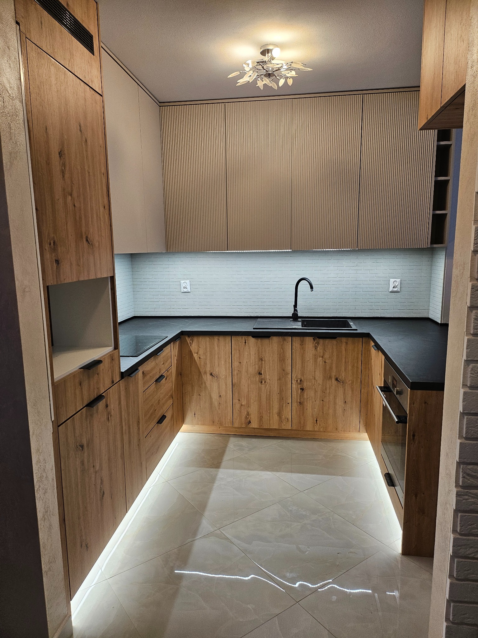 Nowoczesna kuchnia w kształcie litery U z drewnianymi frontami, czarnym blatem i białymi płytkami imitującymi cegłę. Podświetlenie LED pod szafkami dodaje elegancji.