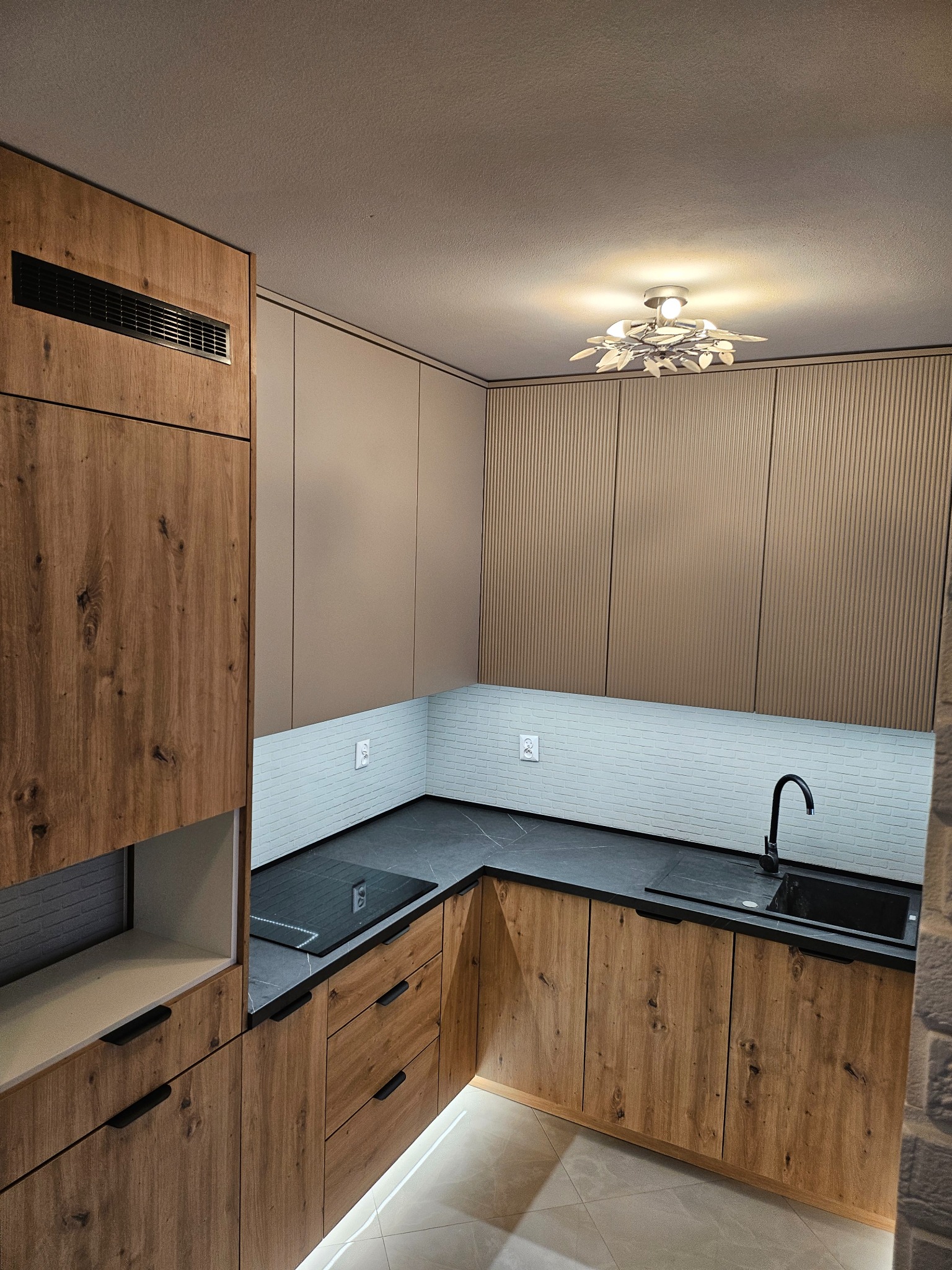 Nowoczesna kuchnia w odcieniach brązu i beżu z drewnianymi frontami, czarnym blatem i subtelnym oświetleniem LED pod szafkami. Uwagę zwraca dekoracyjna lampa sufitowa.