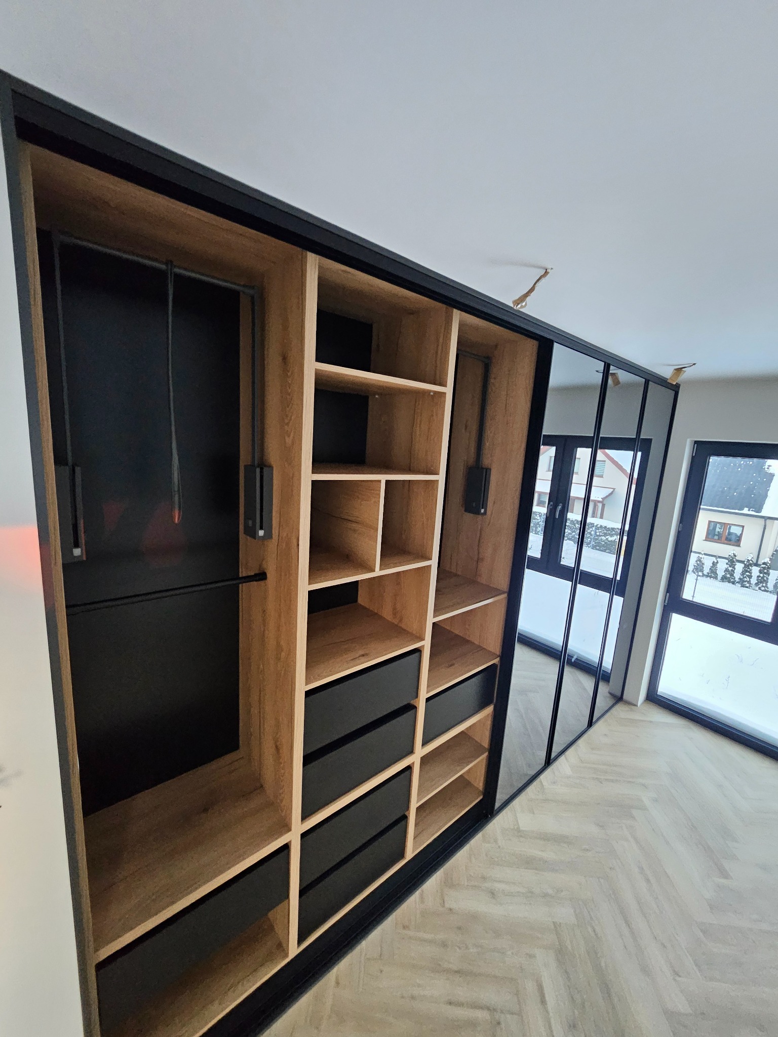 Nowoczesna szafa wnękowa z przesuwnymi lustrzanymi drzwiami i drewnianymi półkami, czarne akcenty, widok na zimowy krajobraz za oknem, parkiet w jodełkę.
