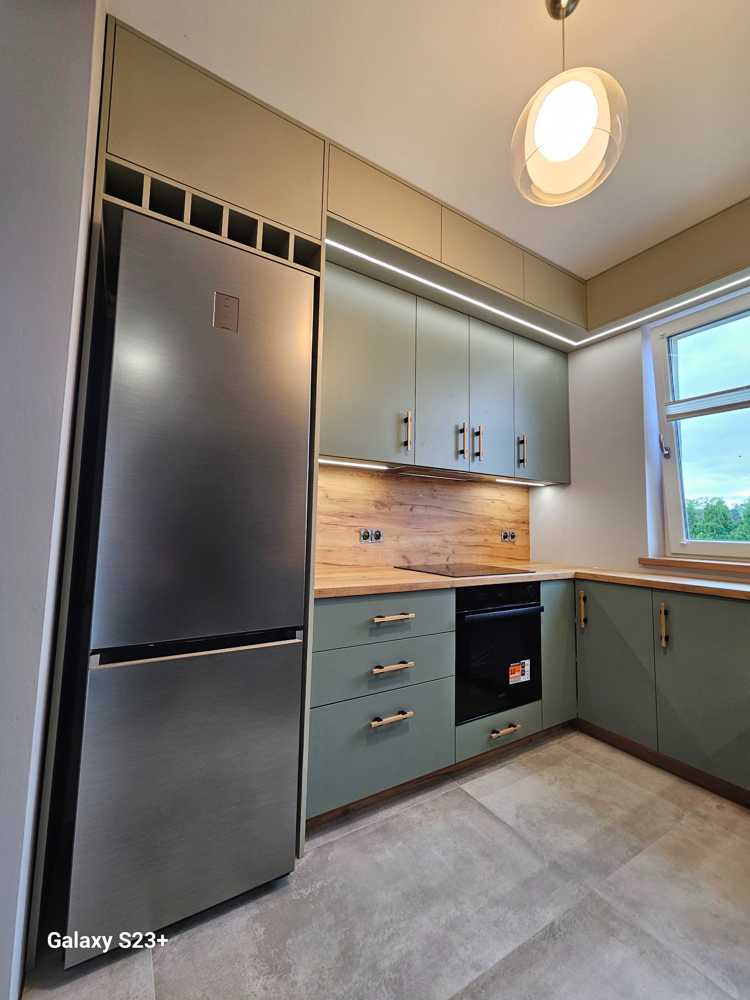 Nowoczesna kuchnia w odcieniach zieleni z drewnianym blatem i srebrną lodówką. Oświetlenie LED podkreśla minimalistyczny design. Widok na okno z zielenią w tle.