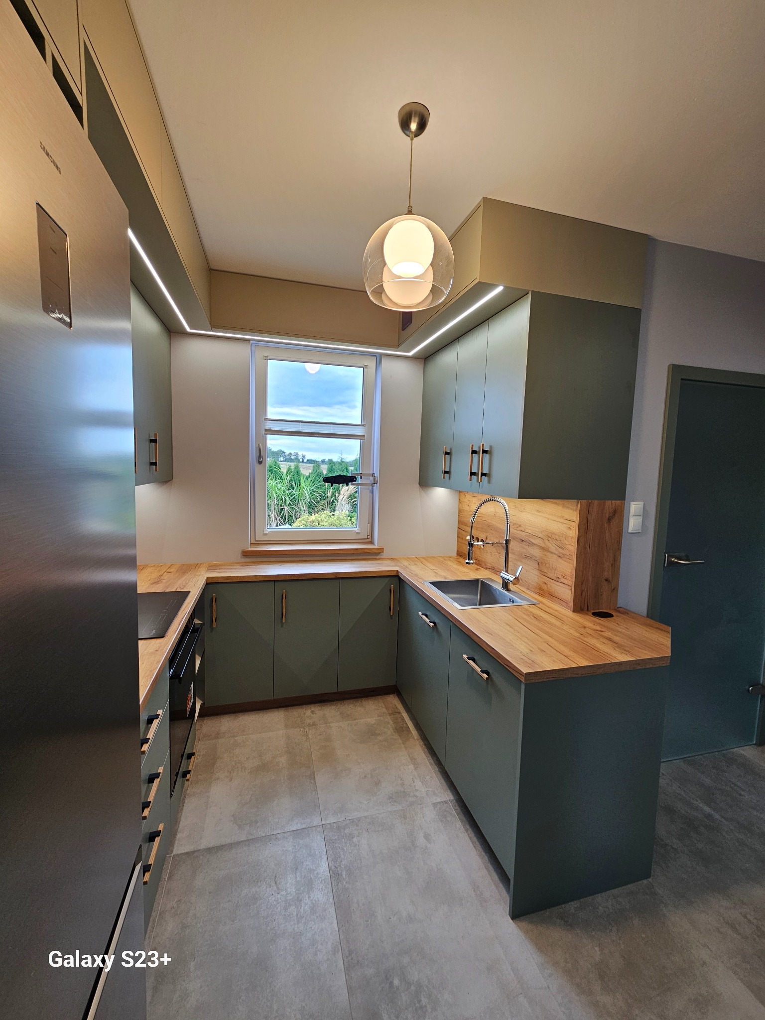 Nowoczesna kuchnia w odcieniach zieleni z drewnianym blatem i oświetleniem LED podszafkowym. Widok na okno z zielenią. Minimalistyczny design z wiszącą lampą.