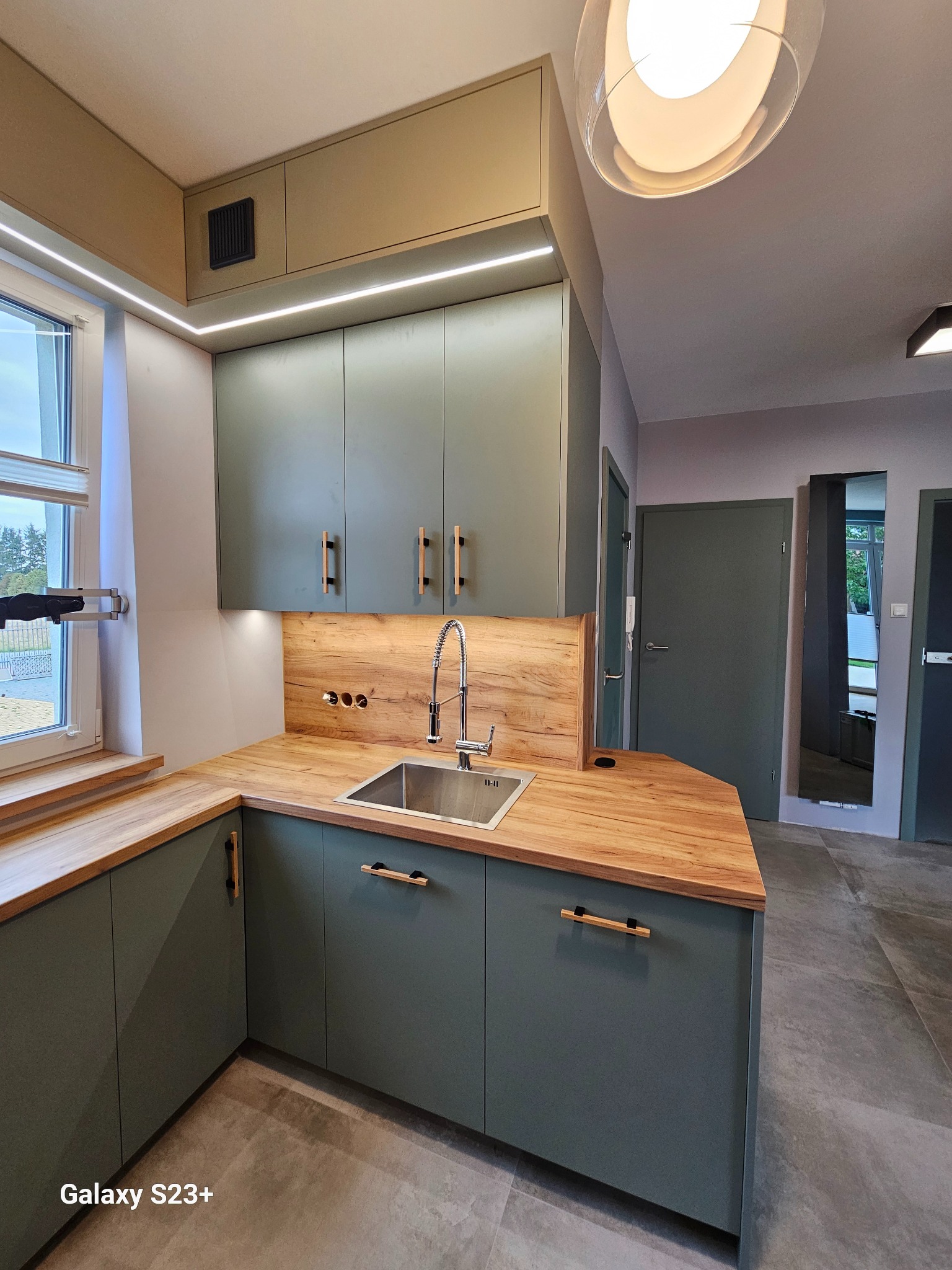 Kompaktowa kuchnia w odcieniach zieleni z drewnianym blatem i oświetleniem LED. Ujęcie z perspektywy, ukazujące detale wykończenia i funkcjonalność zabudowy.