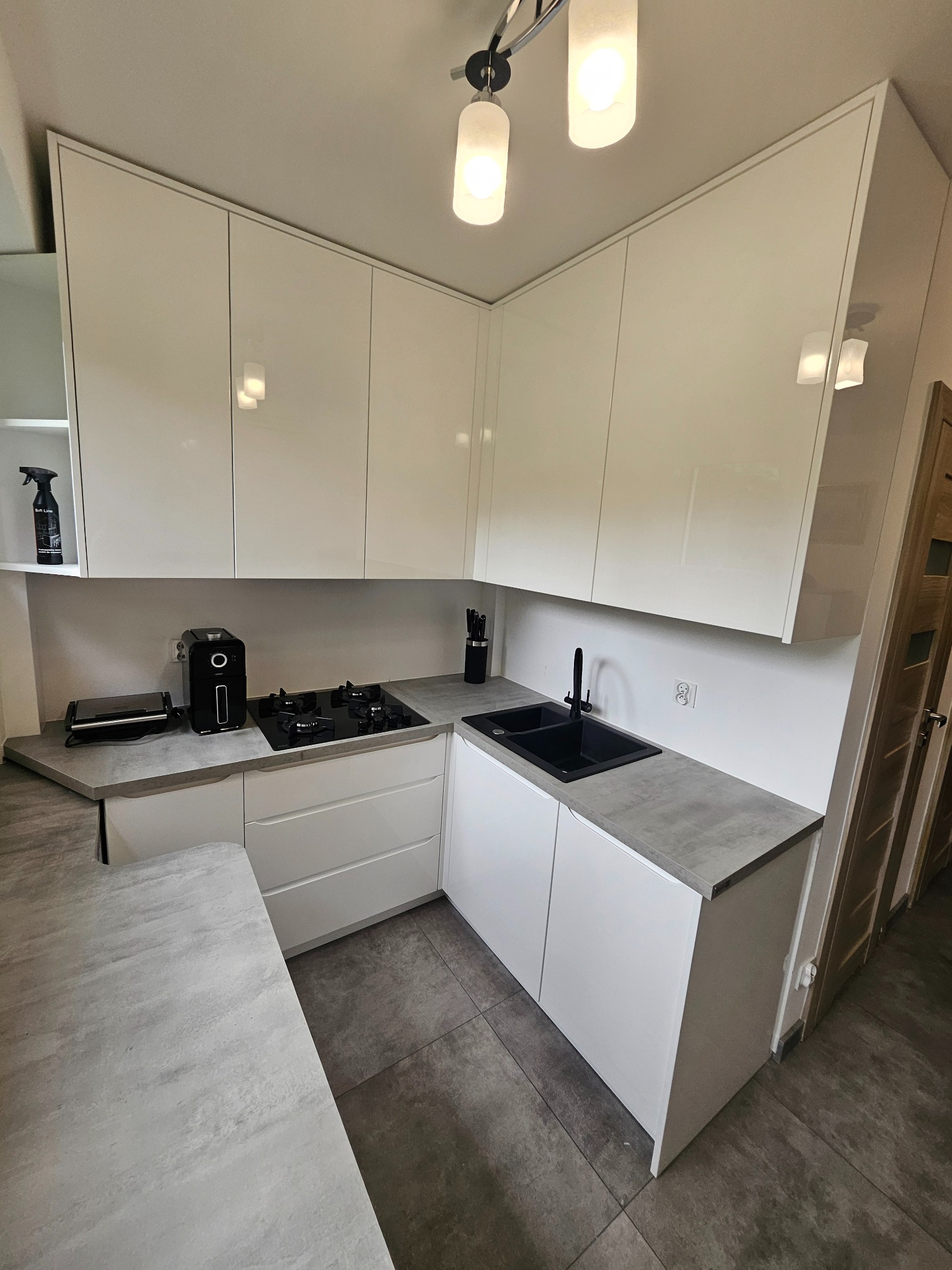 Nowoczesna, biała kuchnia na wymiar z szarym blatem i czarną armaturą. Widoczne sprzęty AGD i oświetlenie sufitowe. Minimalistyczny design.