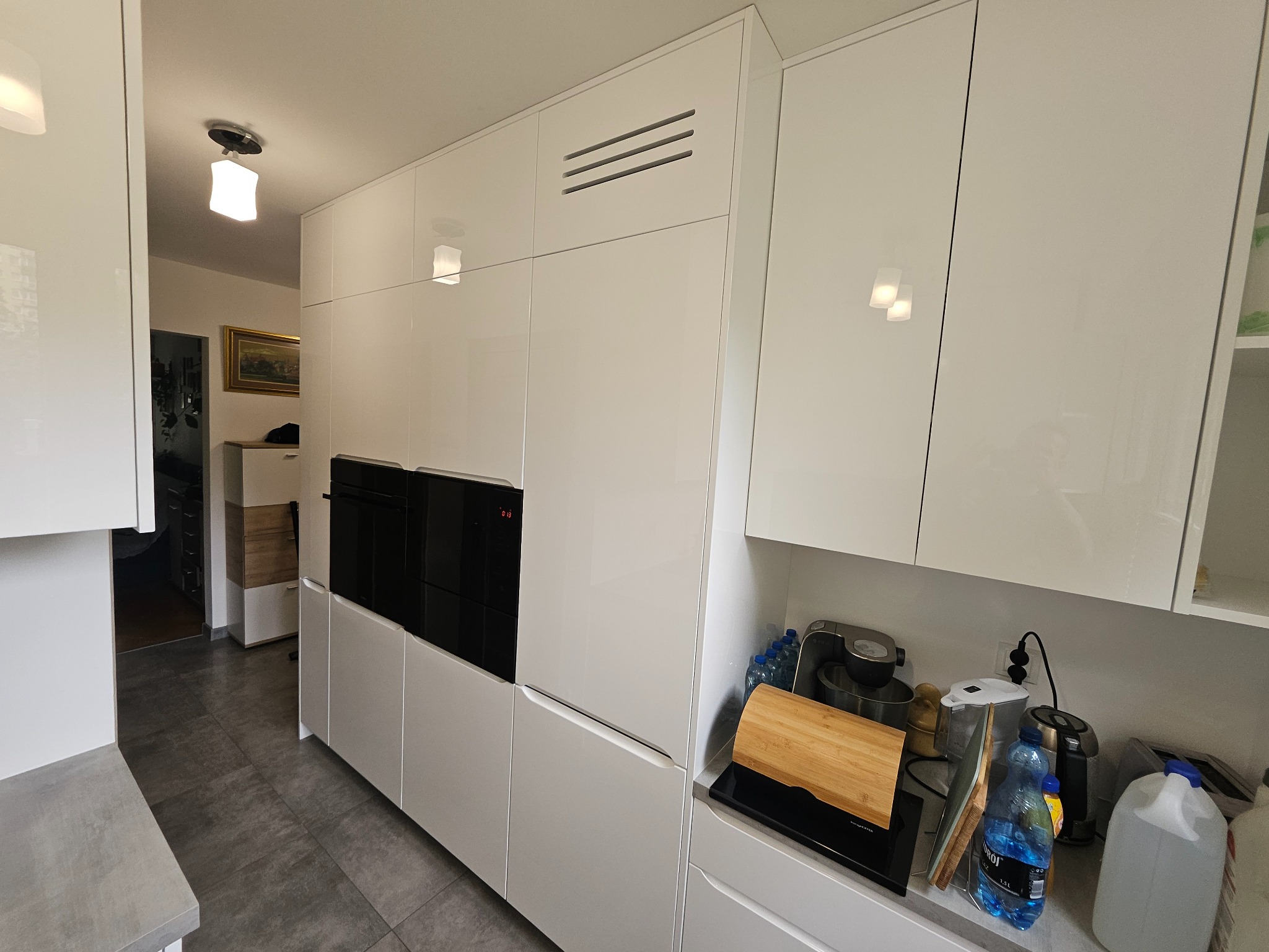 Biała, nowoczesna zabudowa kuchenna z piekarnikiem i szafkami pod sufit. Minimalistyczny design, szare płytki podłogowe, widok na dalszą część mieszkania.