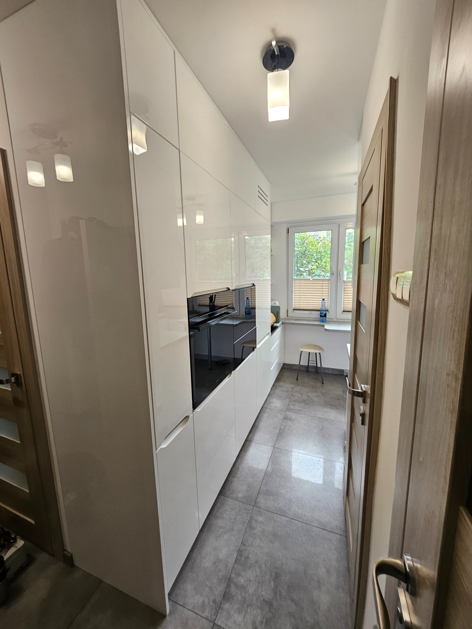 Biała, nowoczesna zabudowa kuchenna z piekarnikiem w zabudowie, szare płytki podłogowe, widok na okno z roletami i krzesłem. Minimalistyczny design.