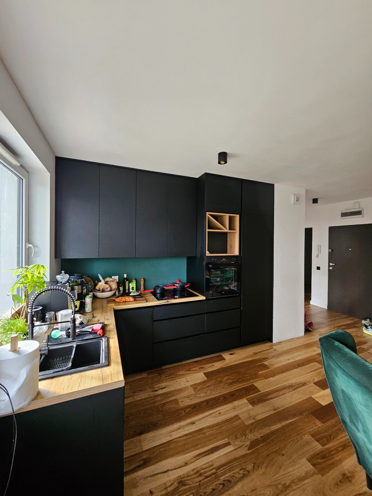 Nowoczesna kuchnia z czarnymi frontami i drewnianym blatem. Widoczne wyposażenie, w tym zlew, płyta grzewcza i piekarnik. Uwagę przykuwa zielona ściana nad blatem.