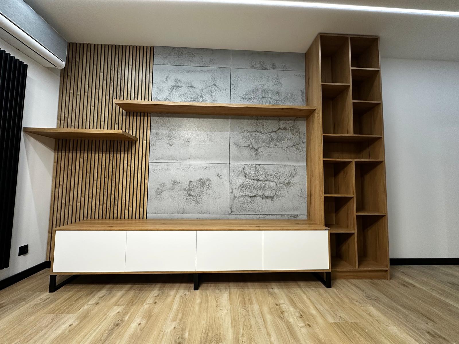 Nowoczesna zabudowa ściany z drewnianymi lamelami, betonowymi panelami i szafką RTV z białymi frontami, uzupełniona o otwarte półki i regał z drewna.