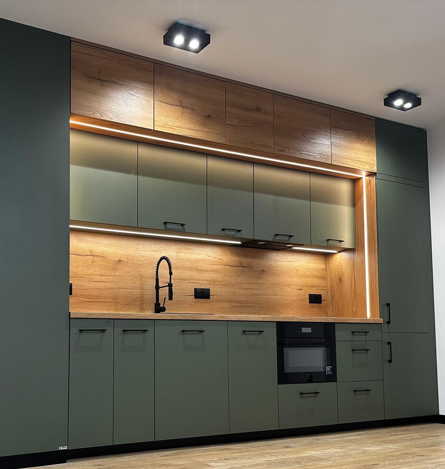Nowoczesna kuchnia z zielonymi szafkami i drewnianymi akcentami, oświetlona listwami LED. Czarna armatura i sprzęt AGD dopełniają elegancki design.