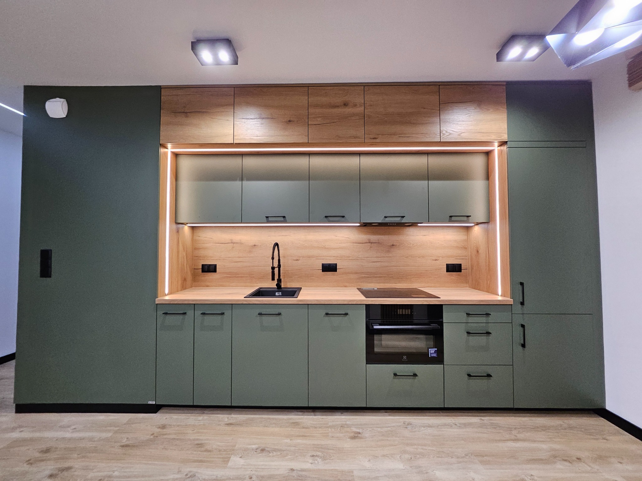 Nowoczesna kuchnia w odcieniach zieleni i drewna z subtelnym oświetleniem LED, minimalistyczne uchwyty, czarny zlew i płyta indukcyjna. Fronty matowe.