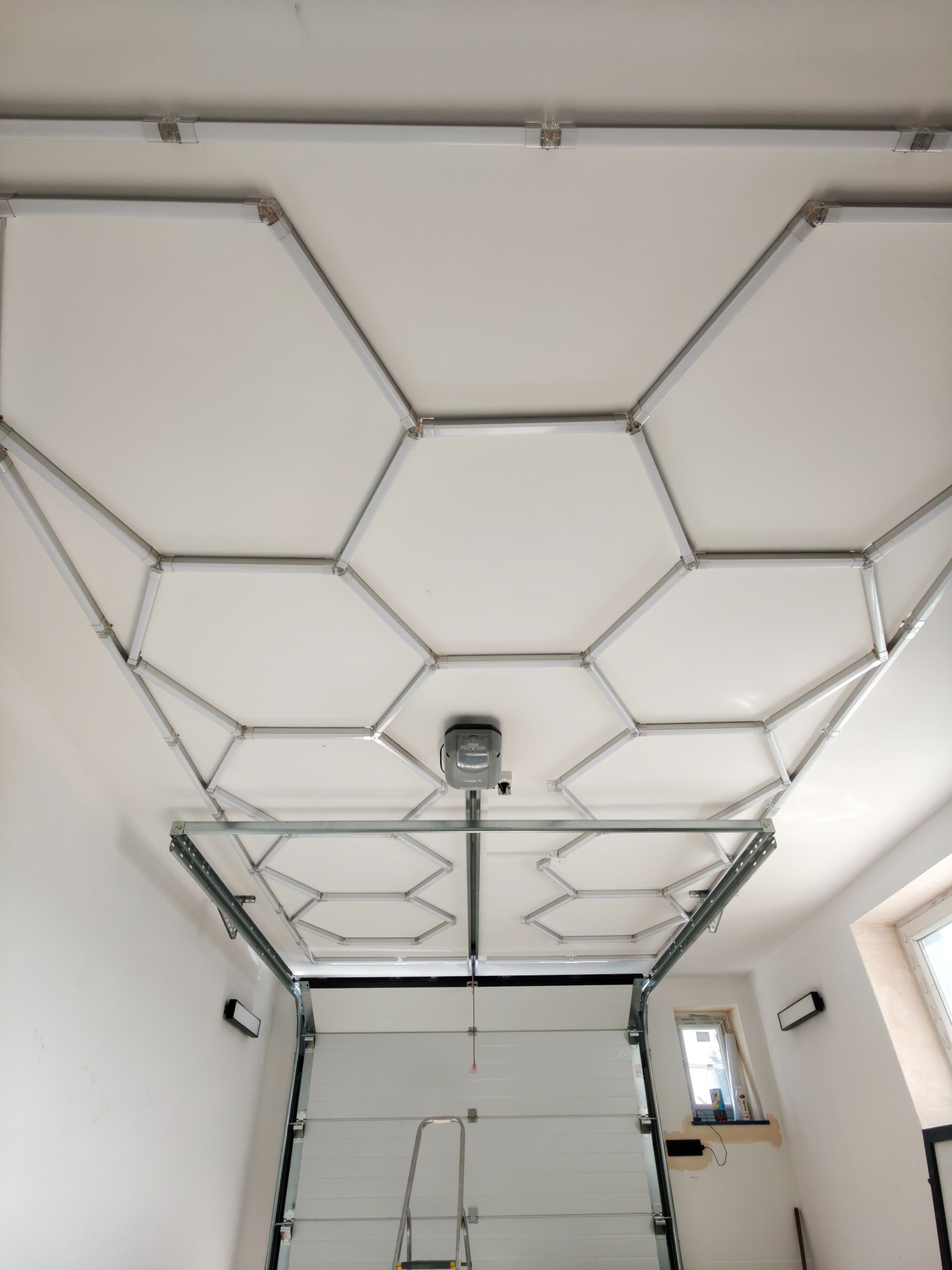 Biała brama garażowa z automatyką, widok od wewnątrz. Na suficie zamontowana konstrukcja z profili, tworząca geometryczny wzór. Widoczna drabina i okno.