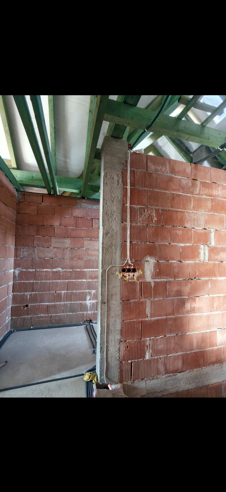 Wnętrze w trakcie budowy z widoczną instalacją elektryczną w ścianie z czerwonej cegły. Widoczne kable i puszki elektryczne. Surowy stan wykończenia, zielona konstrukcja dachu.