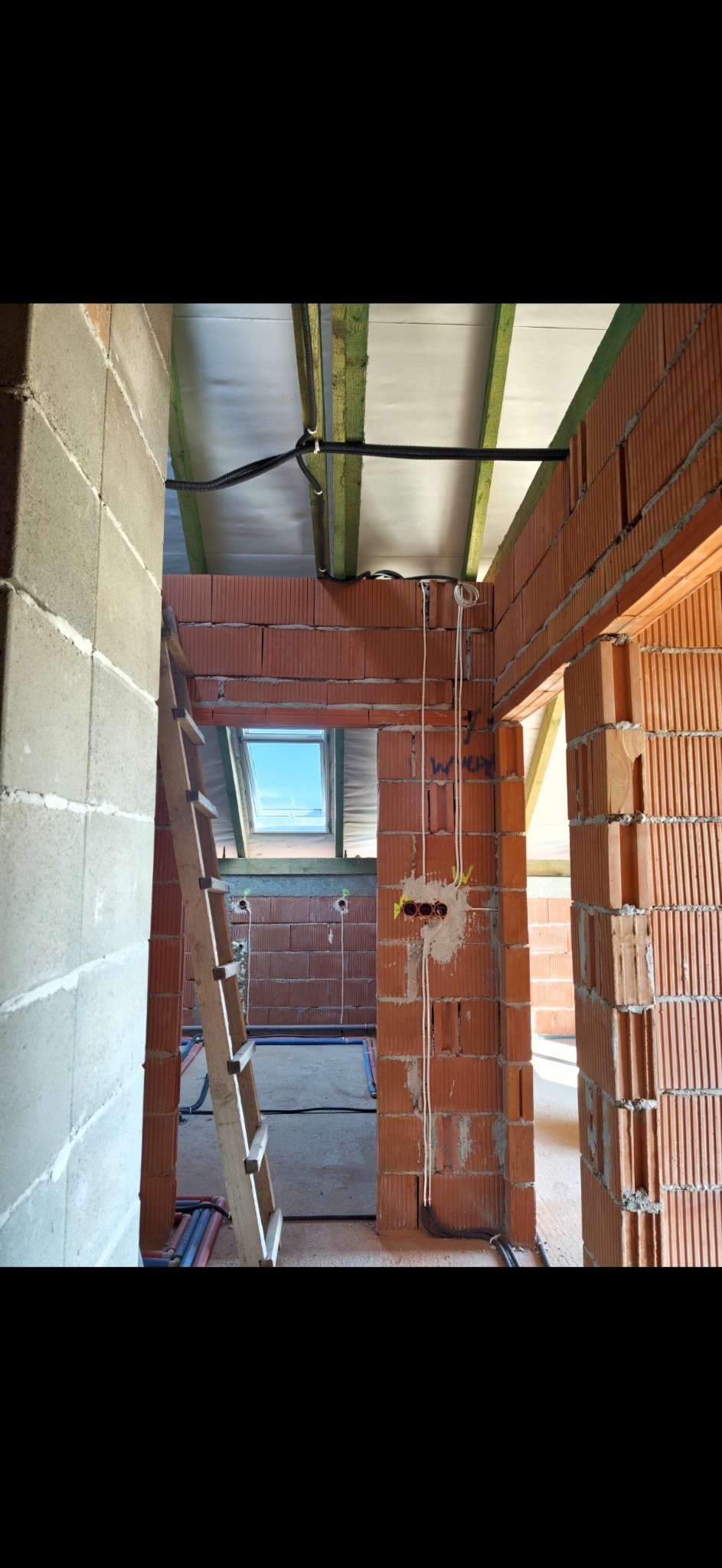 Wnętrze w trakcie budowy z widocznymi przewodami elektrycznymi w ścianach z cegły, drabina oparta o ścianę, okno dachowe w tle. Widoczne kable pod sufitem.