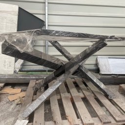 Adam walczy WELLSPAW - Metalowa konstrukcja stołu z krzyżowymi nogami, zabezpieczona folią, leży na palecie w magazynie. Elementy metalowe w kolorze czarnym.