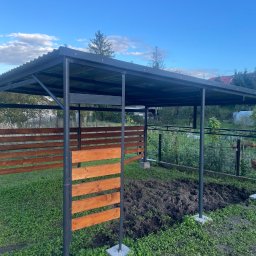 Adam walczy WELLSPAW - Metalowa konstrukcja z drewnianymi panelami w ogrodzie, prawdopodobnie wiata lub pergola. Konstrukcja pomalowana na czarno, panele drewniane w ciepłym odcieniu brązu.