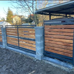 Adam walczy WELLSPAW - Nowoczesne ogrodzenie z drewnianymi panelami i metalową ramą, osadzone na betonowych słupkach. Widoczne elementy konstrukcyjne i surowe wykończenie terenu.