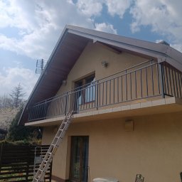 Adam walczy WELLSPAW - Dom z balkonem zabezpieczonym metalową balustradą w kolorze grafitowym. Do balkonu przystawiona drabina. Widok z dołu na budynek w słoneczny dzień.