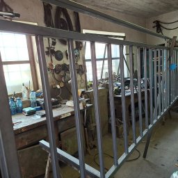 Adam walczy WELLSPAW - Surowa, metalowa konstrukcja balustrady w warsztacie, z widocznymi narzędziami i butlą gazową. Ujęcie z bliska, ukazujące detale spawów i niedokończony charakter.