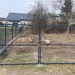 Adam walczy WELLSPAW - Nowoczesne, grafitowe ogrodzenie panelowe z bramą dwuskrzydłową na tle domu jednorodzinnego i drzew bez liści. Widoczne palety i elementy montażowe na posesji.