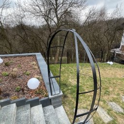 Adam walczy WELLSPAW - Metalowa, czarna pergola ogrodowa w kształcie łuku, zamontowana na kamiennych schodach w ogrodzie. W tle drzewa bez liści i fragment budynku.