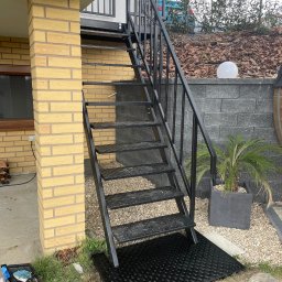 Adam walczy WELLSPAW - Zewnętrzne schody metalowe, czarne, prowadzące na balkon. Widoczne detale konstrukcyjne i otoczenie: ceglana kolumna, rośliny ozdobne, kamień dekoracyjny.
