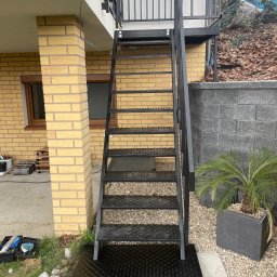 Adam walczy WELLSPAW - Zewnętrzne schody metalowe z balustradą, prowadzące na balkon. Konstrukcja pomalowana na czarno, z perforowanymi stopniami. Obok roślina w donicy i narzędzia.