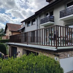 Adam walczy WELLSPAW - Nowoczesna, ciemnobrązowa balustrada tarasowa z pionowych profili na tle jasnej elewacji domu, z dwoma mniejszymi balkonami z ozdobnymi balustradami powyżej. W tle zielone krzewy i zachmurzone niebo.