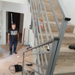 Adam walczy WELLSPAW - Szara, geometryczna balustrada schodowa z metalu, zamontowana przy schodach wyłożonych płytkami imitującymi drewno. W tle widać mężczyznę i narzędzia, co sugeruje etap prac wykończeniowych.