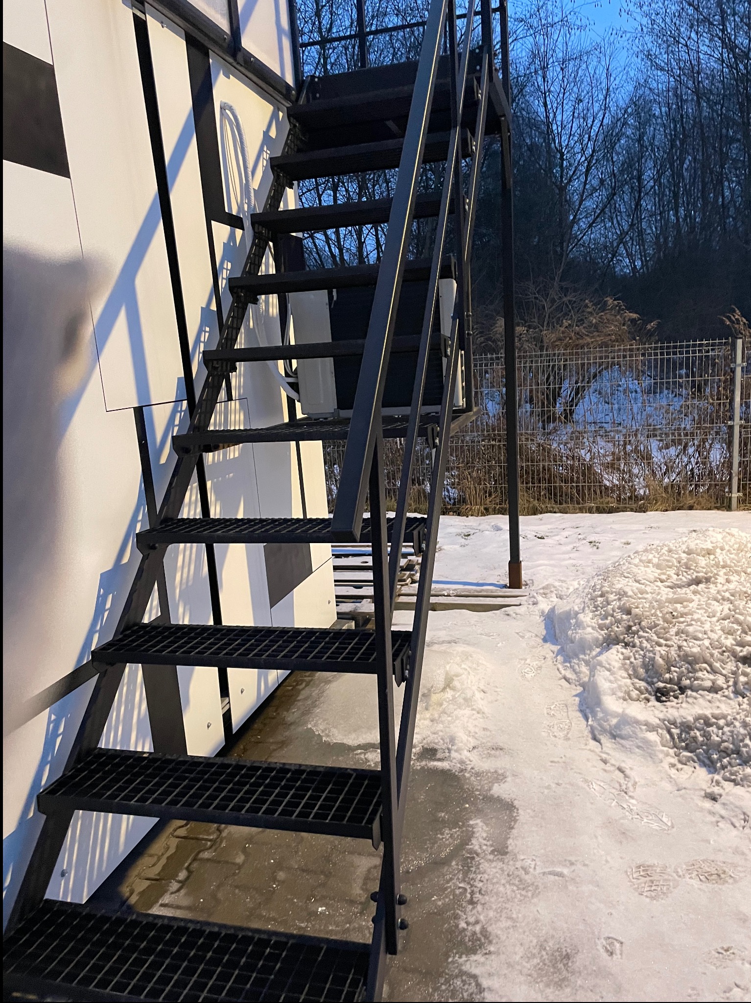 Zewnętrzne schody metalowe z kratowymi stopniami, prowadzące do budynku o biało-czarnej fasadzie. W tle widoczne drzewa i ogrodzenie w zimowej scenerii.