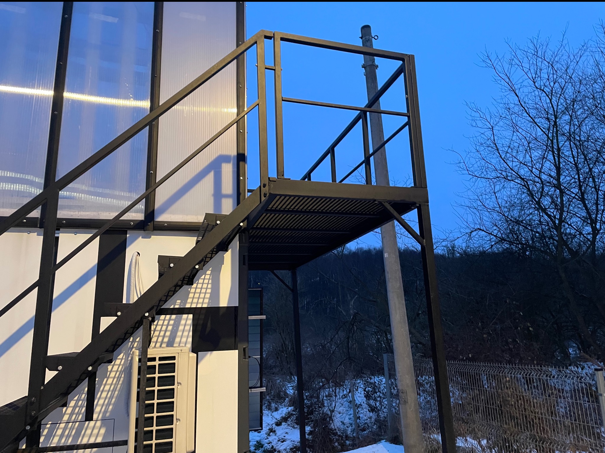 Zewnętrzne, metalowe schody z podestem, prowadzące do budynku o nowoczesnej architekturze. Konstrukcja pomalowana na czarno, widoczne detale łączeń. Tło: zimowy krajobraz.