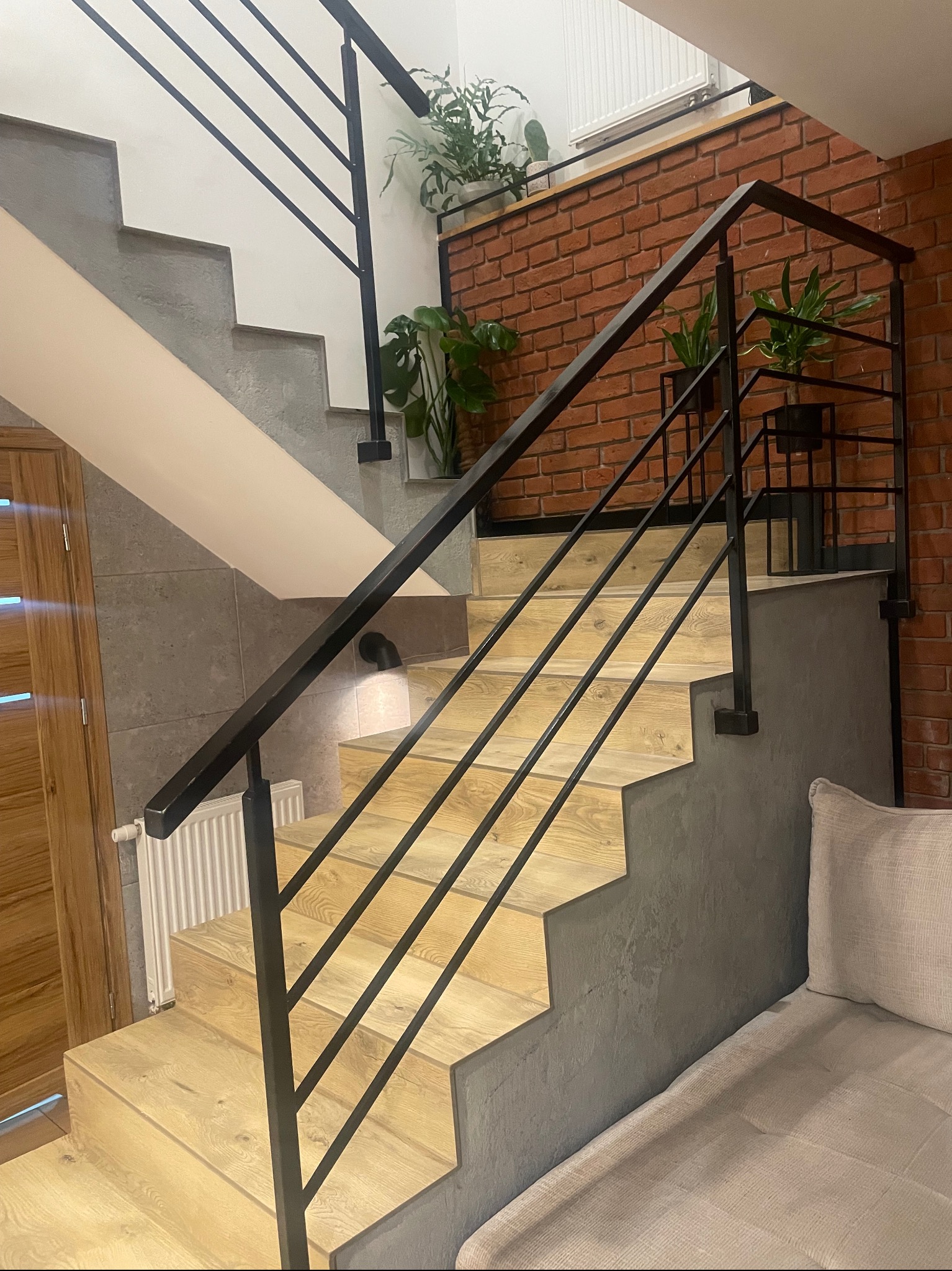 Nowoczesne schody z drewnianymi stopniami i czarną, metalową poręczą na tle ceglanej ściany i roślin doniczkowych. Minimalistyczny design z akcentami natury.